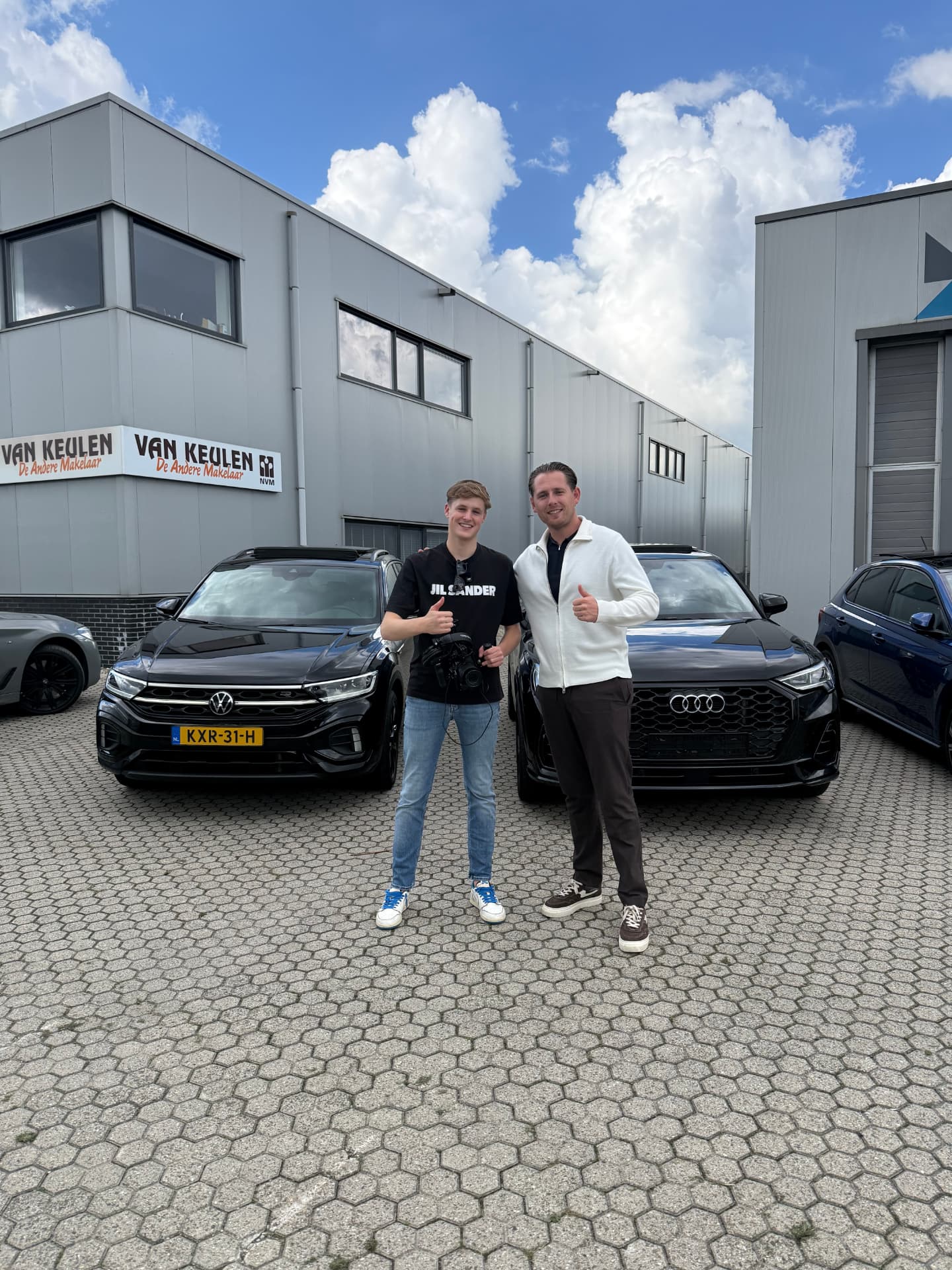 Twee mannen poseren met duim omhoog tussen twee auto's na de opnames
